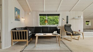 A modern living room with a black leather sofa, a wooden coffee table, and a black armchair. A large window provides a view of greenery outside. - Beach front home in Rudkøbing with sauna (Rudkøbing)