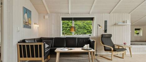Een moderne woonkamer met een zwarte leren bank, een houten salontafel en een zwarte fauteuil. Een groot raam biedt uitzicht op groen buiten.