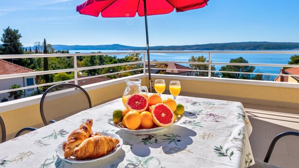 A balcony with a table set for breakfast, including croissants, oranges, and a pitcher of juice, overlooking a scenic view of the sea and hills.
