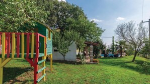Una casetta gioco verde con uno scivolo, un piccolo edificio bianco con un pilastro in mattoni e una zona piscina con lettini.