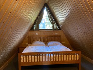 Ein Zimmer mit schräg stehender Decke, Holzwänden, einem Bett mit Holzrahmen und einem Fenster mit Vorhängen.