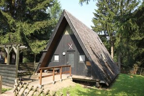 Eine dreieckige Holzhütte mit Tür und Fenstern, umgeben von Bäumen.