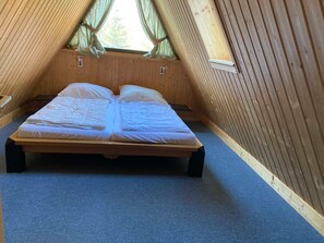 A single bed in a wooden room with a sloped ceiling and a window with curtains.