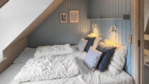 Ein Schlafzimmer mit schrägem Dach, einem Bett mit weißer Bettwäsche und gemusterten Kissen, zwei gerahmten Pflanzenprints an der Wand und Wandleseleuchten.