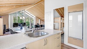 Cocina moderna con una isla grande, un fregadero y armarios de madera. Hay una zona de comedor con una mesa y sillas, y vistas a árboles a través de amplios ventanales.