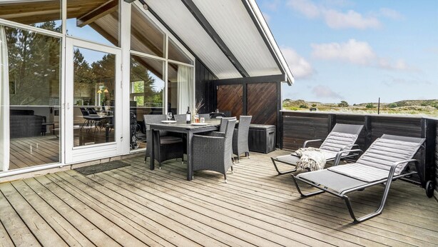 Una terraza de madera con mobiliario exterior, que incluye una mesa y sillas, y un área de descanso con tumbonas.