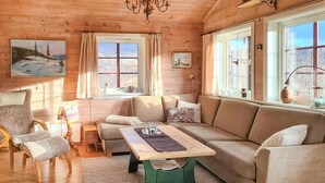 Interior acogedor de una cabaña con piso de madera, una mesa de centro verde, un sofá color beige y una silla. Hay dos ventanas con cortinas, una araña y un cuadro en la pared.