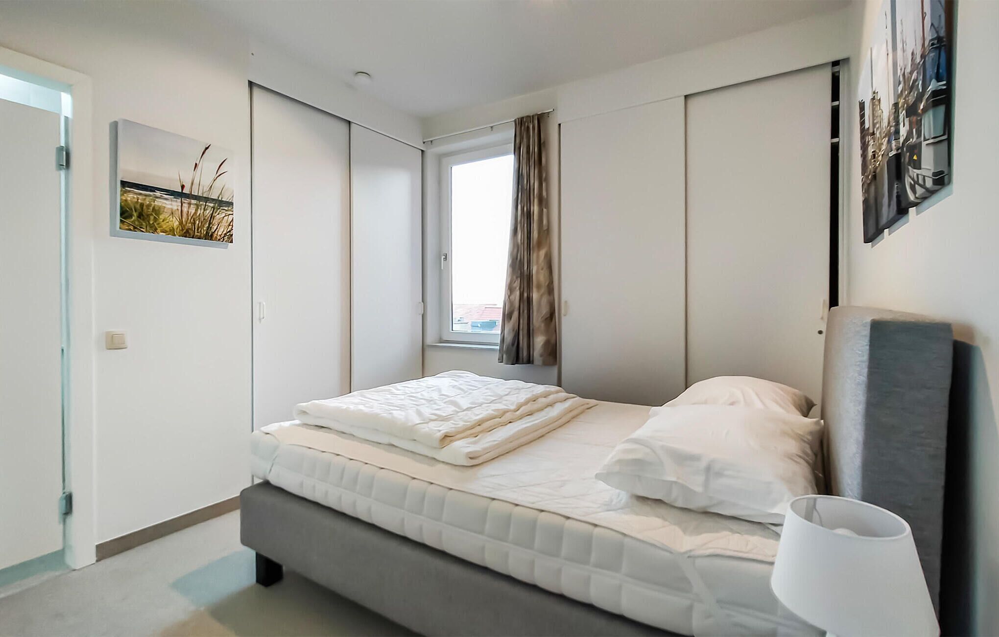 A modern bedroom with a bed, a window with curtains, a painting, and a lamp.