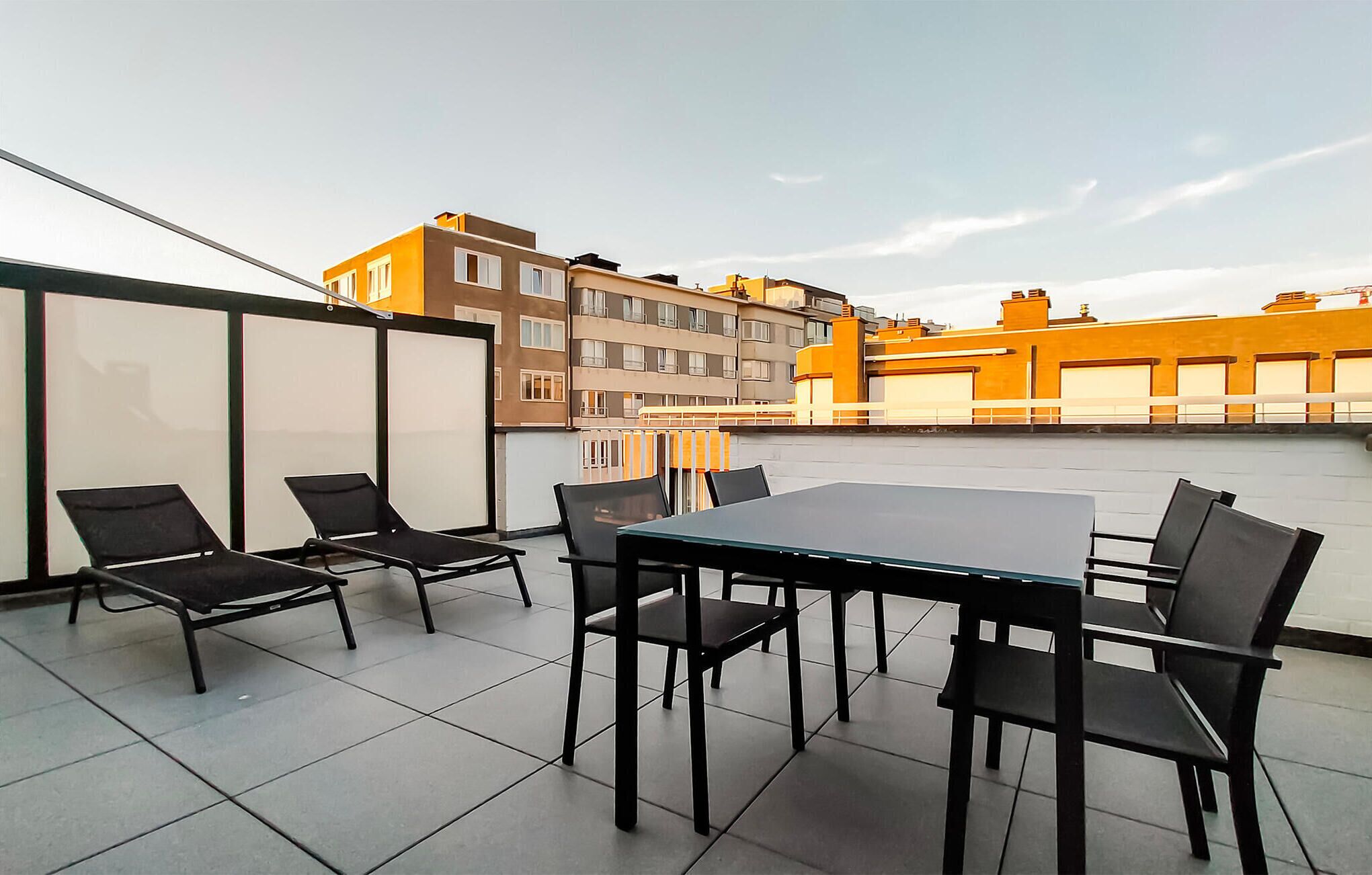Terraço no telhado com mesa e cadeiras, divisória de vidro e vista dos edifícios ao redor.