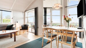 A spacious room with a large dining table, chairs, a sofa, and a TV. There are large windows providing natural light and a view of the outdoors.
