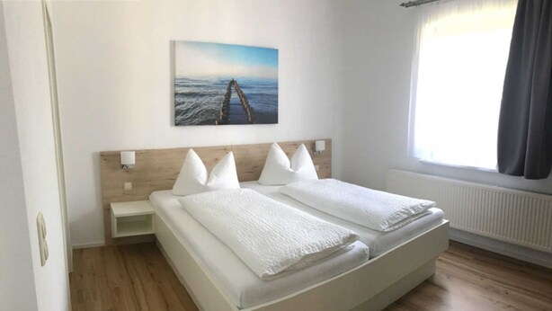 A bedroom with a wooden headboard, a white bed with two pillows, a bedside table with a lamp, and a window with curtains. - Apartment Type 1 - Meutzner Trassenheide, "Villa Seute Deern" (Trassenheide)