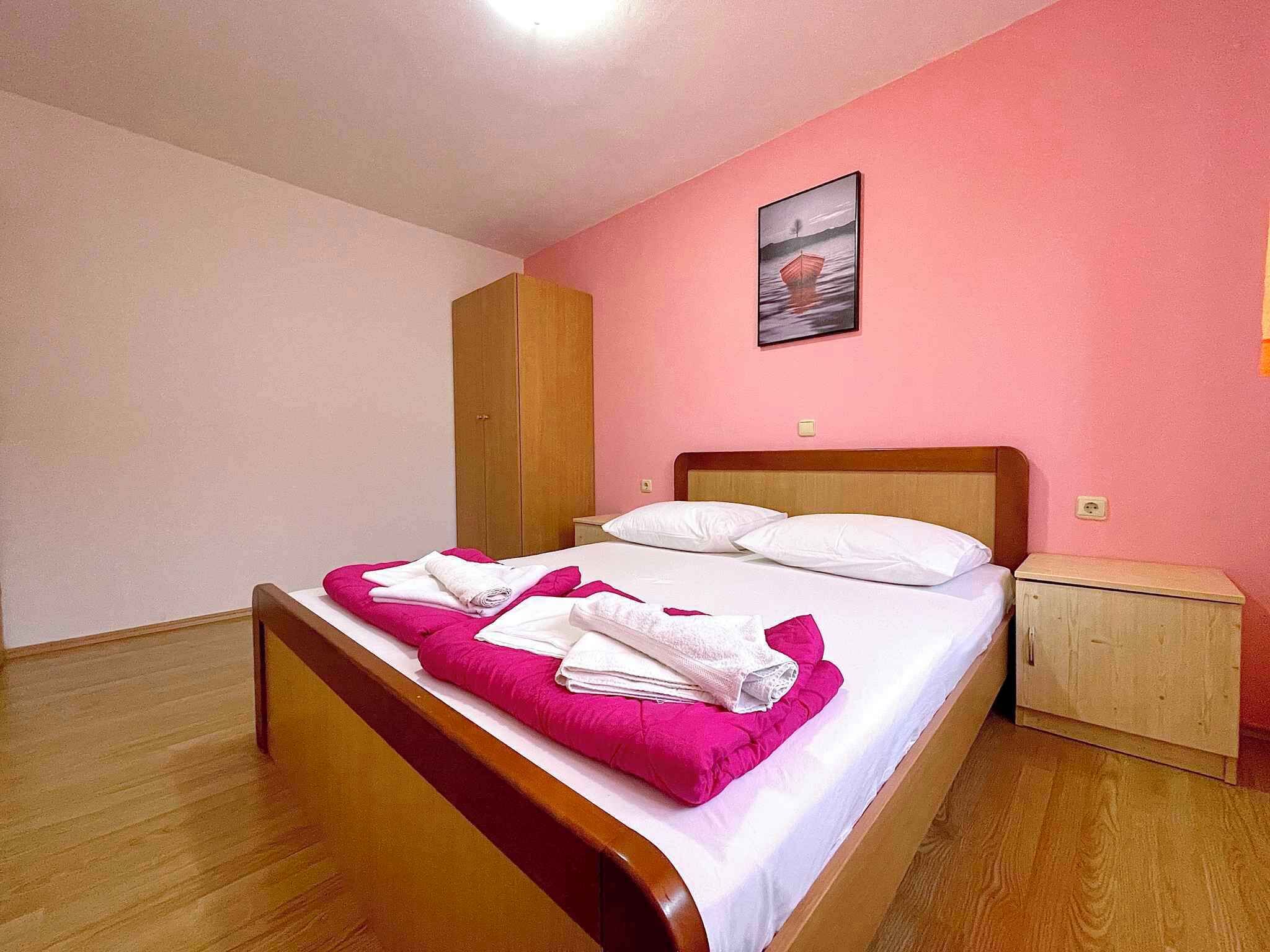 Ein ordentlich bezogenes Bett mit weißen Leinen und rosafarbenen Kissen, ein Holz-Kopfschild, ein Nachttisch und ein Bild an der Wand.