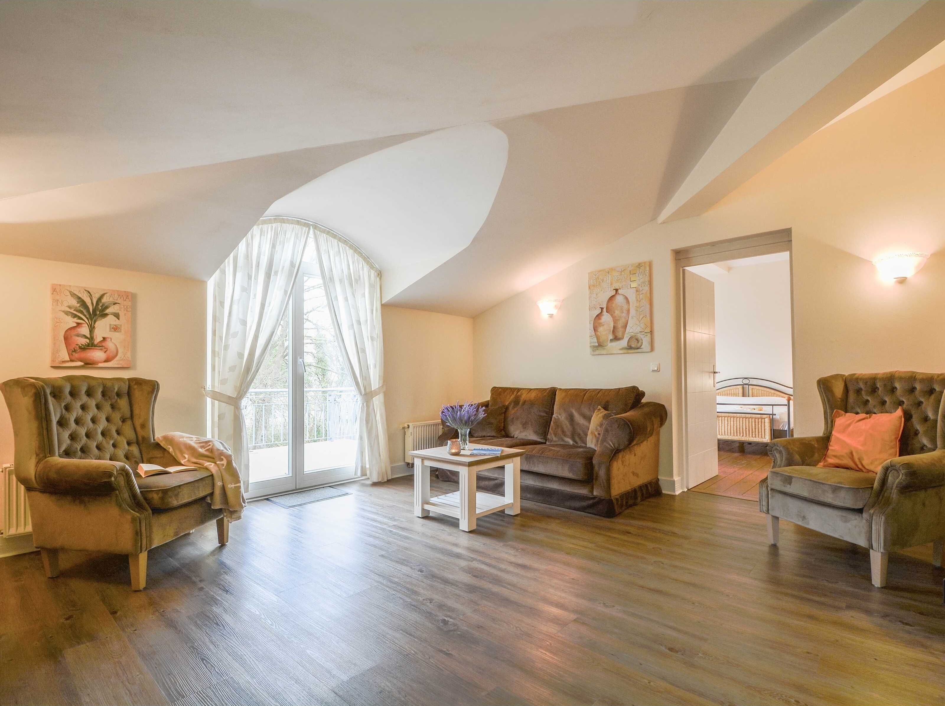 A spacious living room with a large window, a brown sofa, a tufted armchair, and a small white table.