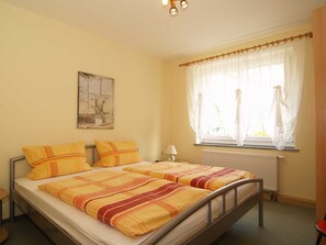 Un lit double avec une literie à rayures, une table de chevet, une lampe, une fenêtre avec des rideaux légers et un radiateur.