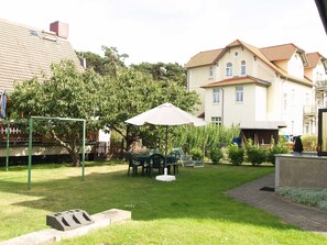 Un jardin à l’arrière d’une maison, avec une pelouse, une balançoire, une table avec des chaises et un grand parasol.