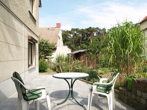 Une terrasse avec une table ronde et deux chaises, entourée d’un jardin où poussent différentes plantes et délimitée par une clôture.