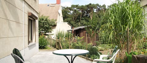 Un patio con una mesa redonda y dos sillas, rodeado de un jardín con diversas plantas y una cerca.