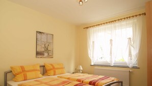 A double bed with striped bedding, a bedside table, a lamp, a window with sheer curtains, and a radiator. - Fritz-Reuter-Str. 10a. Curr. FR-10 - Fritz-Reuter-Str. 10a (Kühlungsborn)