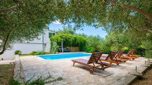 A poolside area with a stone patio, wooden lounge chairs, and a modern building with a glass facade in the background. - Beautiful home in Kastel Sucurac (Kastel Sucurac)