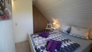 Een slaapkamer met een schuine plafond, een bed met beddengoed met een patroon, een nachtkastje met een lamp, en een stoel.