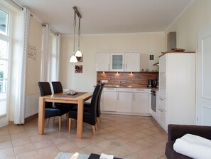 Una cocina moderna con armarios blancos, encimera de madera y horno empotrado. Hay una zona de comedor con una mesa y sillas, y una ventana con cortinas estampadas.