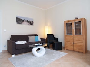 Una sala de estar con un sofá marrón, una mesa de centro de vidrio, un otoman blanco, un sillón negro y un mueble de madera con puertas de cristal.