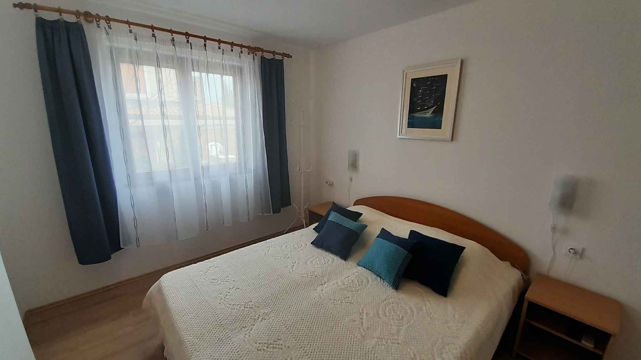 Une chambre à coucher avec un lit, une table de chevet, une fenêtre avec des rideaux et un tableau encadré au mur.