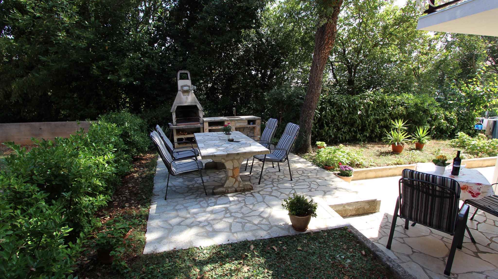 Une terrasse extérieure avec une table et des chaises en pierre, entourée d’une végétation luxuriante et agrémentée d’un barbecue.