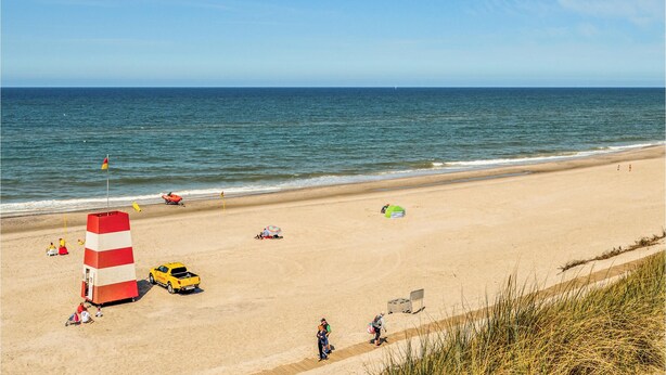 A beach with a lifeguard tower, a yellow vehicle, and people walking along the shore. - Lovely home in Ringkøbing with sauna (Ringkøbing)