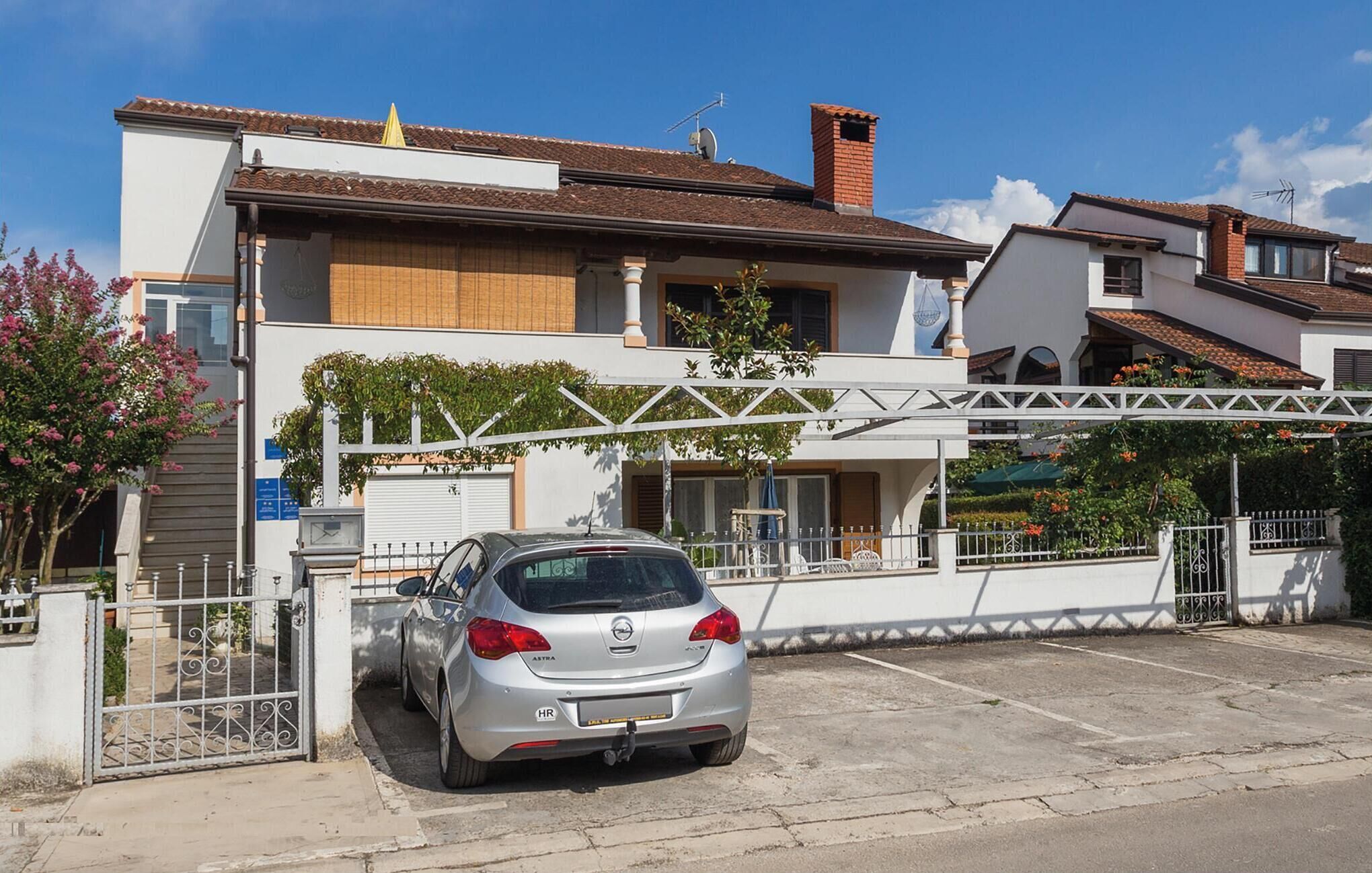 A silver car parked in a gated driveway of a residential building with a white facade and a tiled roof.