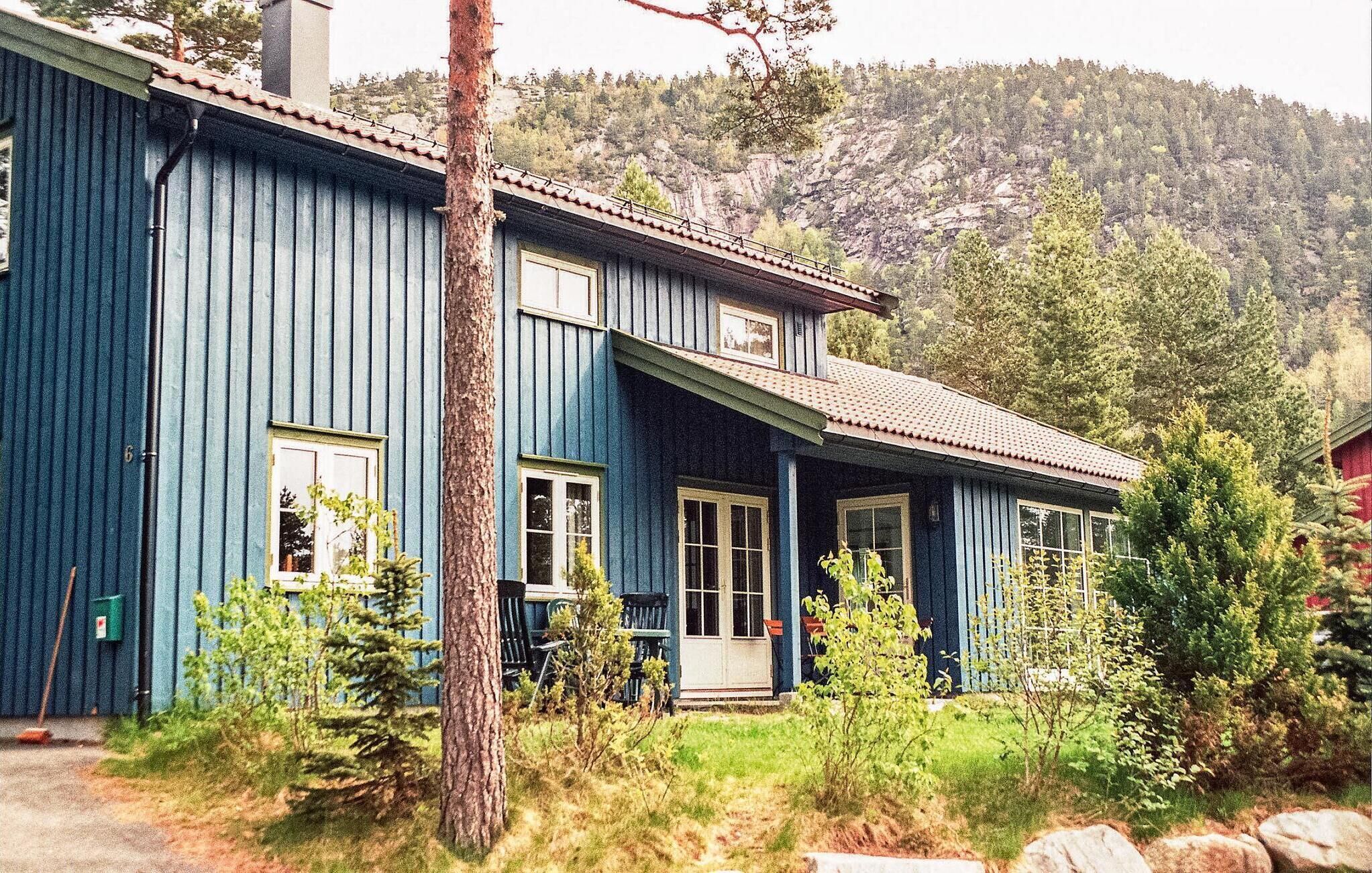 A two-story blue house with a front porch, surrounded by greenery and a mountainous backdrop.
