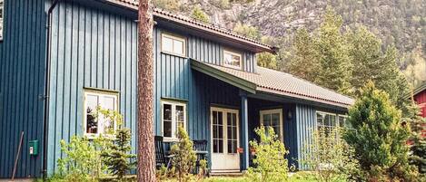 A two-story blue house with a front porch, surrounded by greenery and a mountainous backdrop.