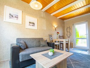 Ein gemütliches Wohnzimmer mit einer grauen Couch, einem Holztisch und einem Essbereich mit Tisch und Stühlen. An der Wand hängen zwei gerahmte Bilder, eine Wandleuchte und ein großes Fenster mit Blick auf Grünfläche.