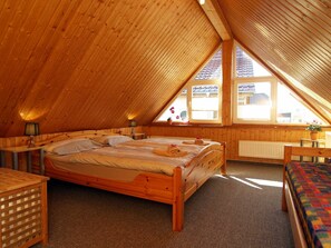 A cozy attic bedroom with a sloped ceiling, wooden walls, a large bed, a bedside table, a chair, and a window with a view of the outside.