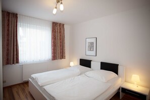 A hotel room with two single beds, a white radiator, a window with curtains, a bedside table with a lamp, and a framed picture on the wall.