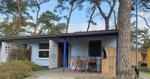 A building with a blue exterior, a door open to a room, a table with chairs, and a window. - Zinno BU 02 - Zinnowitz Pine Route 5c Ferienbungalow 02 (Zinnowitz)