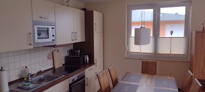 Una cocina con microondas, estufa y lavamanos. Hay una ventana con persianas y una lámpara colgante.