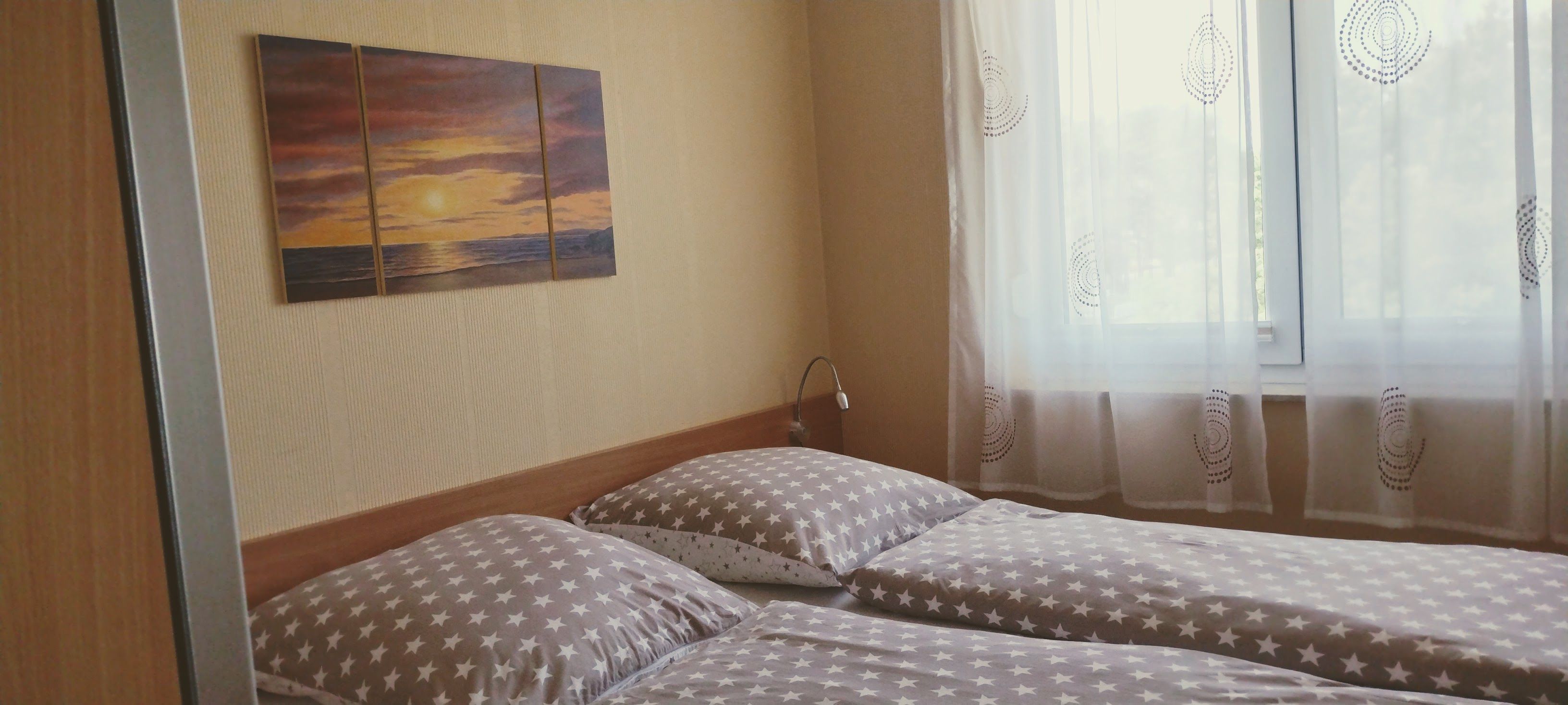 Ein Zimmer mit zwei Betten, einem an der Wand hängenden Gemälde und einem Fenster mit transparenten Vorhängen.