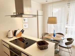 Cozinha moderna com fogão por indução, forno embutido e exaustor. Há uma área de refeições com mesa de vidro e jogos americanos trançados.