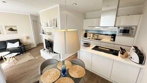 Cocina moderna con armarios blancos, microondas integrado y un lavamanos. Hay un comedor con una mesa de vidrio y posavasos tejidos.