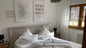 Una cama bien hecha con ropa de cama blanca y varias almohadas, una mesita de noche con lámpara, y una pared decorada con obras enmarcadas.