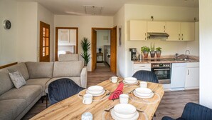 Un salón acogedor con un sofá y una mesa de comedor preparada para cuatro personas. Se ve una cocina al fondo, con armarios blancos y una estufa.