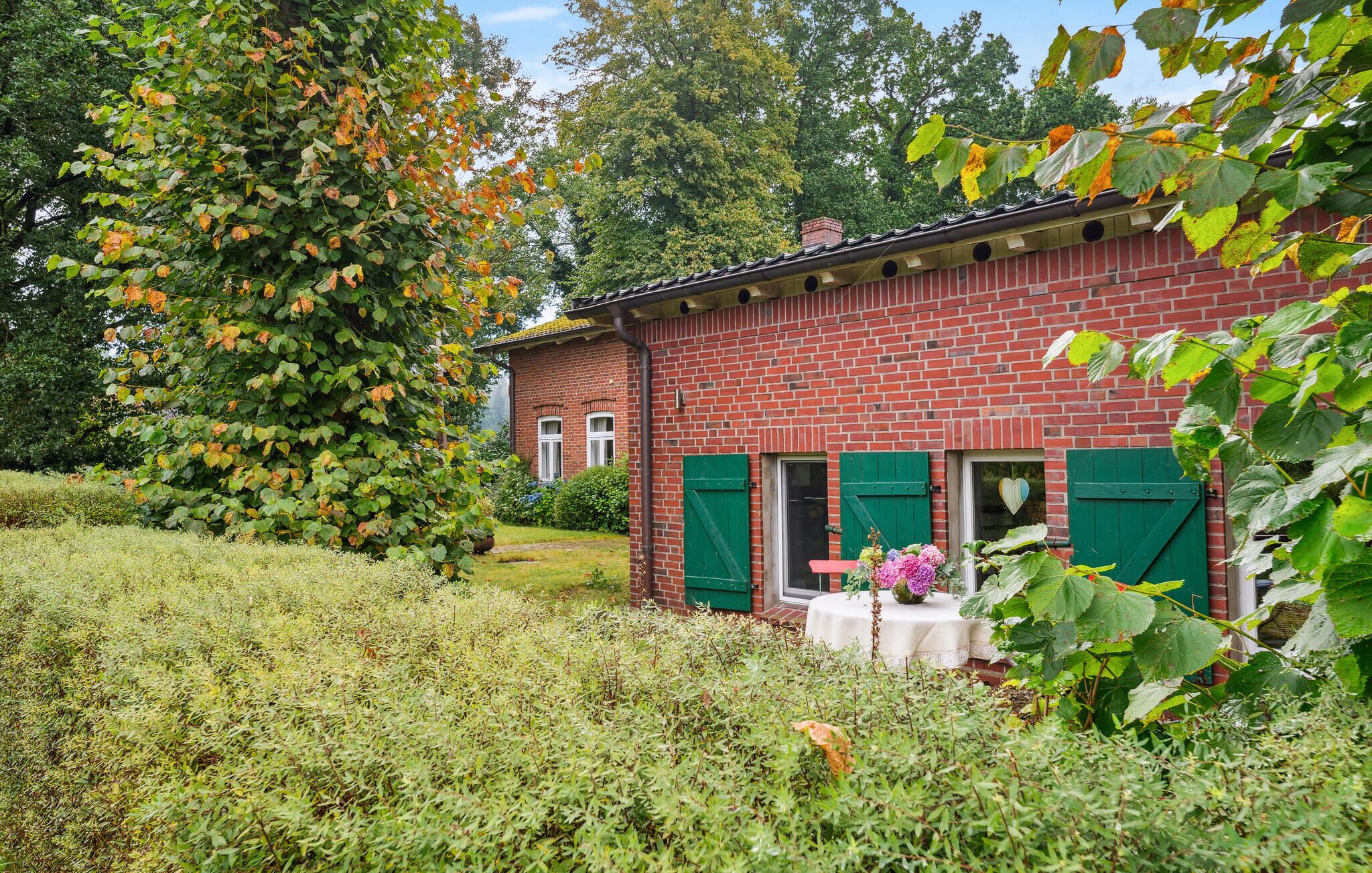 Een rode bakstenen woning met groene luiken, een tuin met diverse planten en een tafel met bloemen.