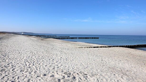 Een strand met een houten pier die in het water uitloopt, zandstrand en een helderblauwe lucht.
