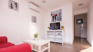 Una sala de estar moderna con un sofá blanco, una mesita con una planta y un televisor sobre un mueble blanco. Se ve una cocina al fondo.