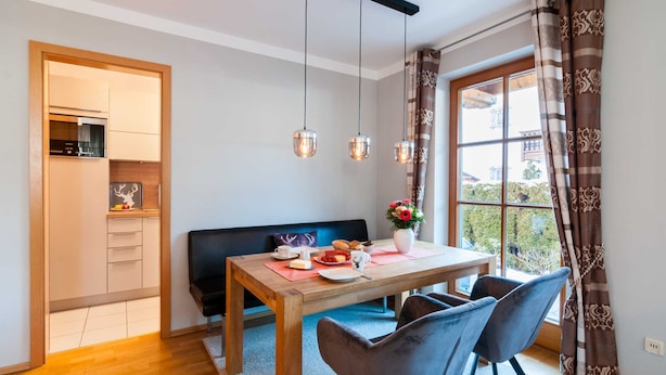 Een eetgedeelte met een houten tafel, twee stoelen, een bank en uitzicht op een balkon door een raam met gordijnen.