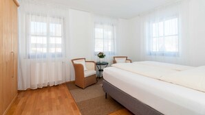 Een hotelkamer met een bed, twee stoelen, een klein tafeltje en een raam met dunne gordijnen.