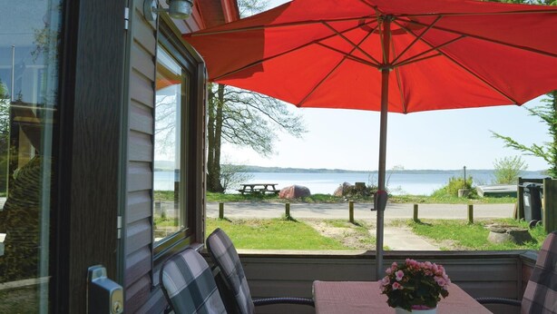 Eine Terrasse mit rotem Sonnenschirm, Sitzgelegenheiten und Blick auf einen See.