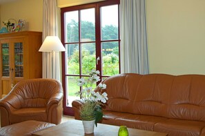 A room with a brown leather sofa, a wooden cabinet, a table with a vase of flowers, and a large window with curtains.