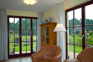 A room with a large sliding glass door leading to a garden, a leather chair, a wooden cabinet, and a table with chairs outside.